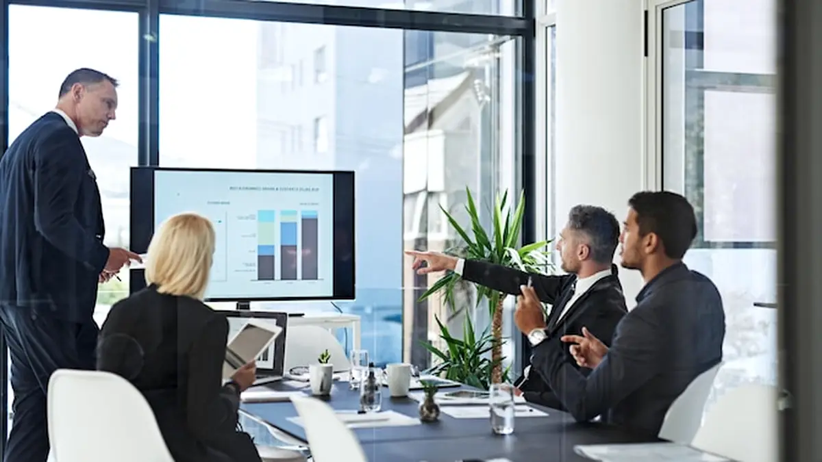 People around a boardroom table while a man in a dark suit presents a bar chart on a wall screen