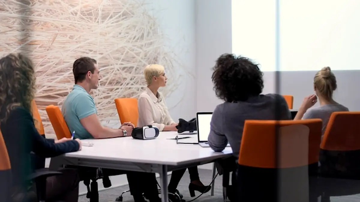 Colleagues seated around a white table in a meeting room with orange chairs and a textured wall
