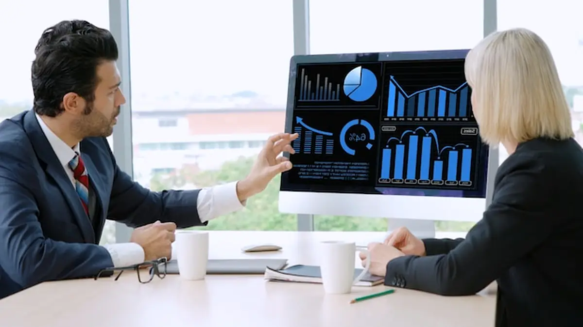 A man in a suit pointing at bar and pie charts on a desktop monitor while a woman looks on