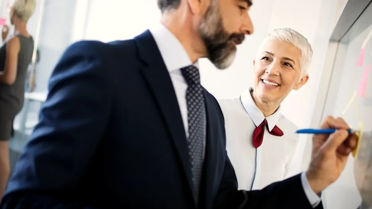 A bearded man in a suit writing on a whiteboard with sticky notes beside a smiling woman